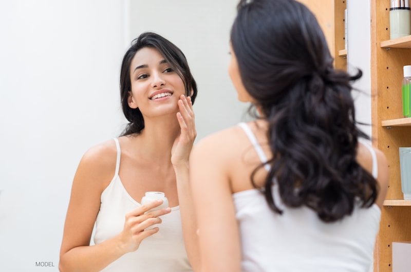 A beautiful Hispanic woman is applying moisturizer to her face while looking in the mirror.